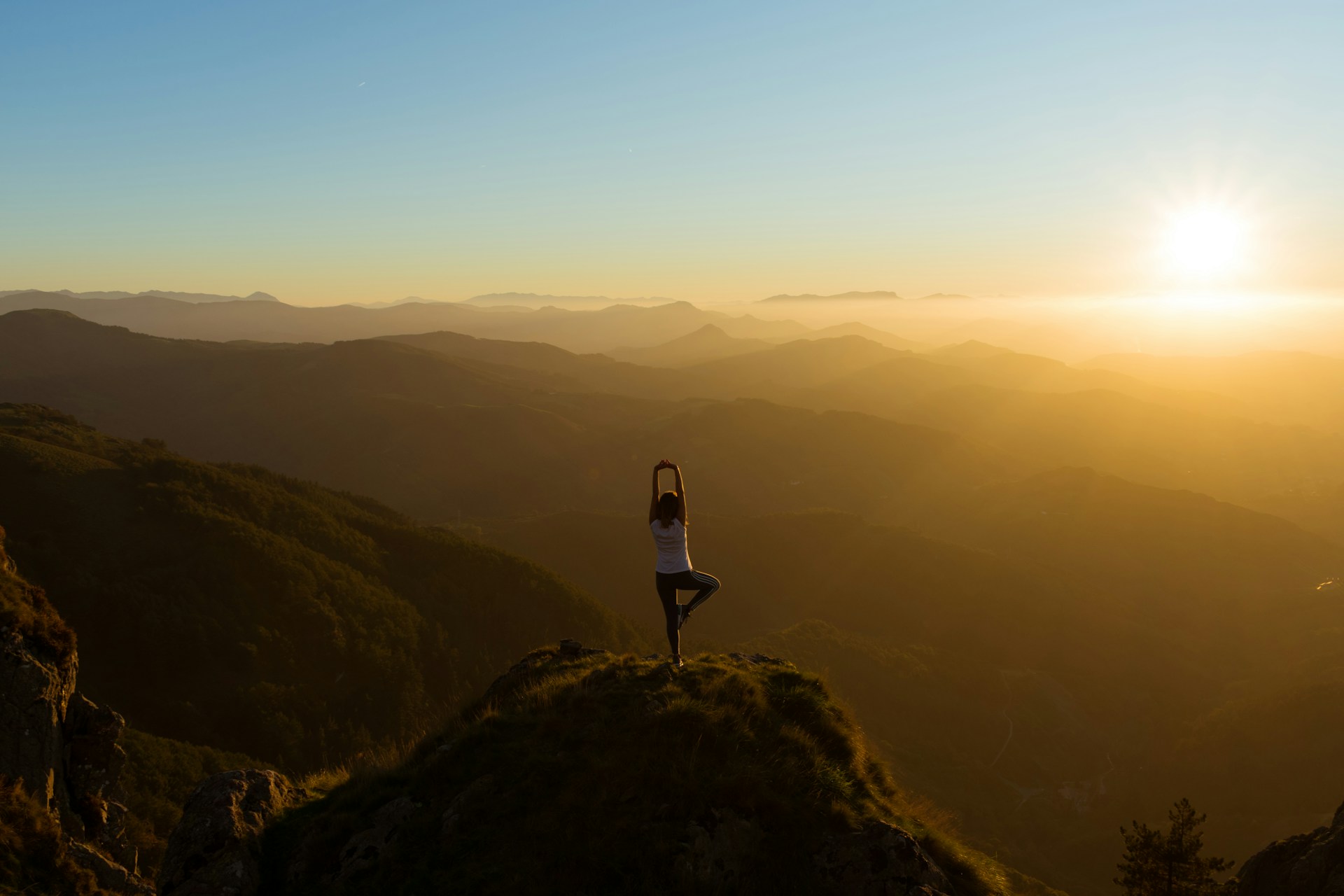 A woman standing on one leg in a yoga pose overlooks a valley at sunset